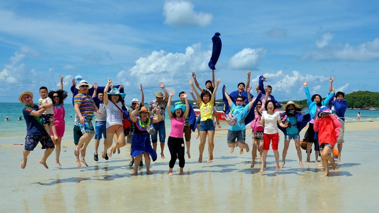 A tour group of 30 travelers jumping for joy in the air on a beach in the sun